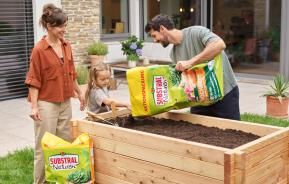 Mutter, Tochter und Vater befüllen ihr Hochbeet mit unserer Substral® Naturen® Hochbeeterde Torffrei.